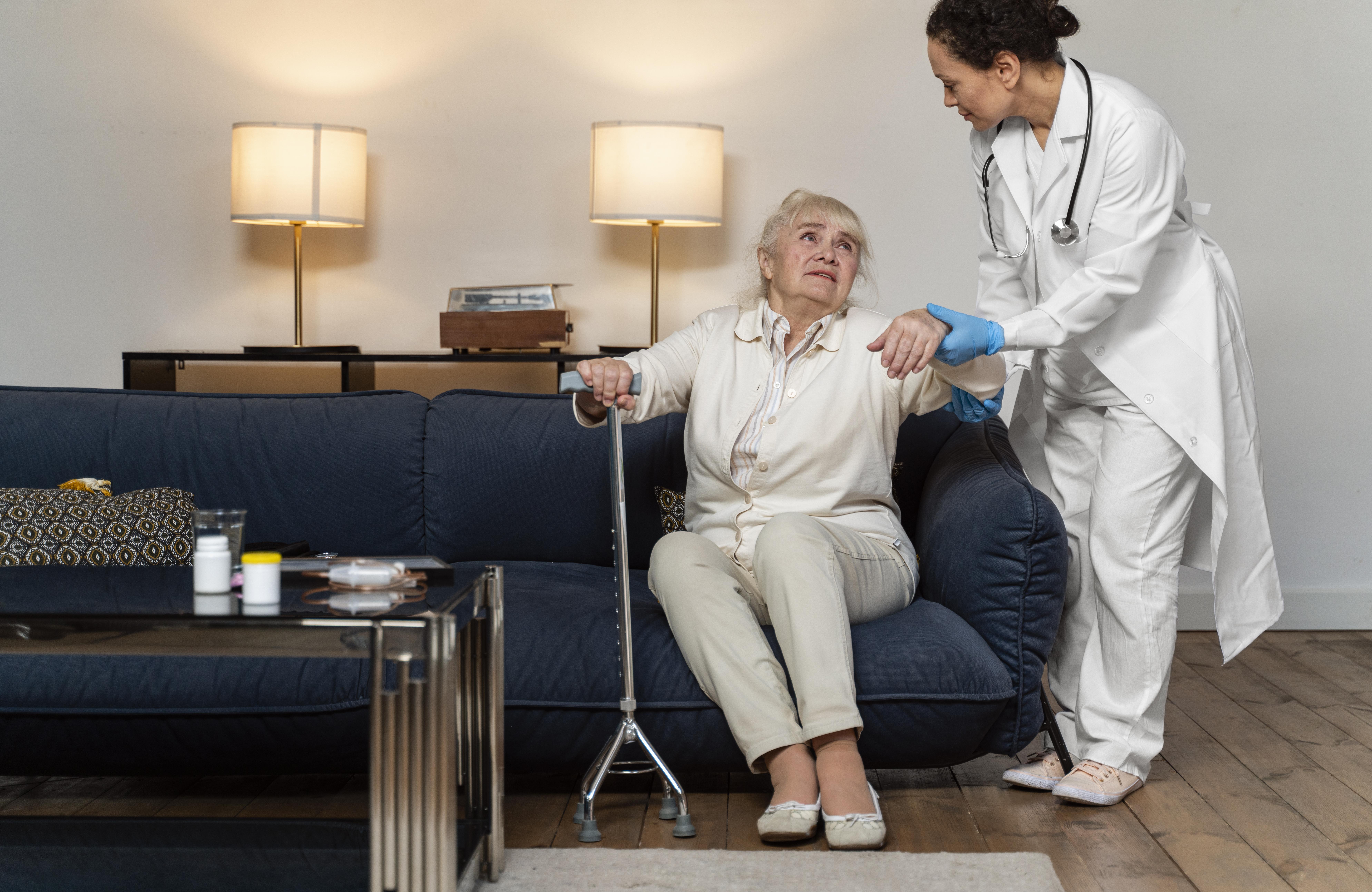 A professional female doctor at home in The Sustainable City helping an elderly patient stand up from a sofa.